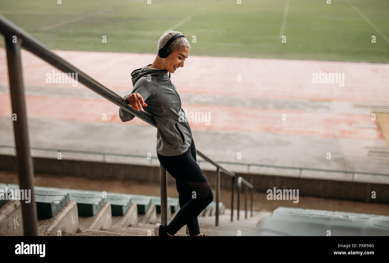 Lächelnde Frau in Fitness Kleidung stand auf der Treppe ein Stadion Musik zu hören. Frau, die auf der Treppe, die zur Unterstützung der Geländer Stockfoto