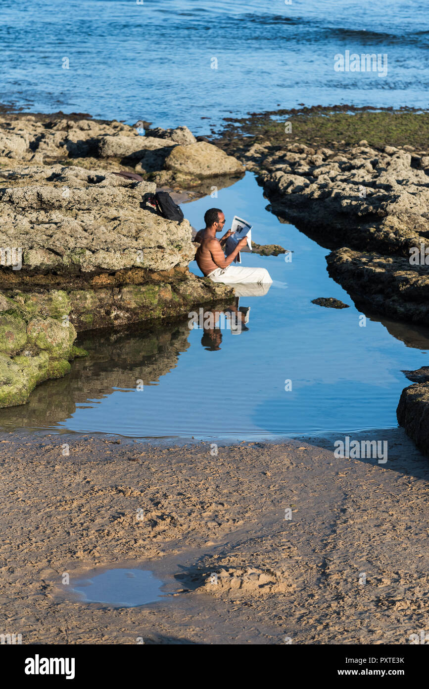 Estoril, Portugal - Juli 9, 2018: der Mann, der in Leinen Hose sitzen in Wassergefüllten rock Pool, der von den atlantischen Meer Lesen einer Zeitung am Nachmittag sunsh Stockfoto