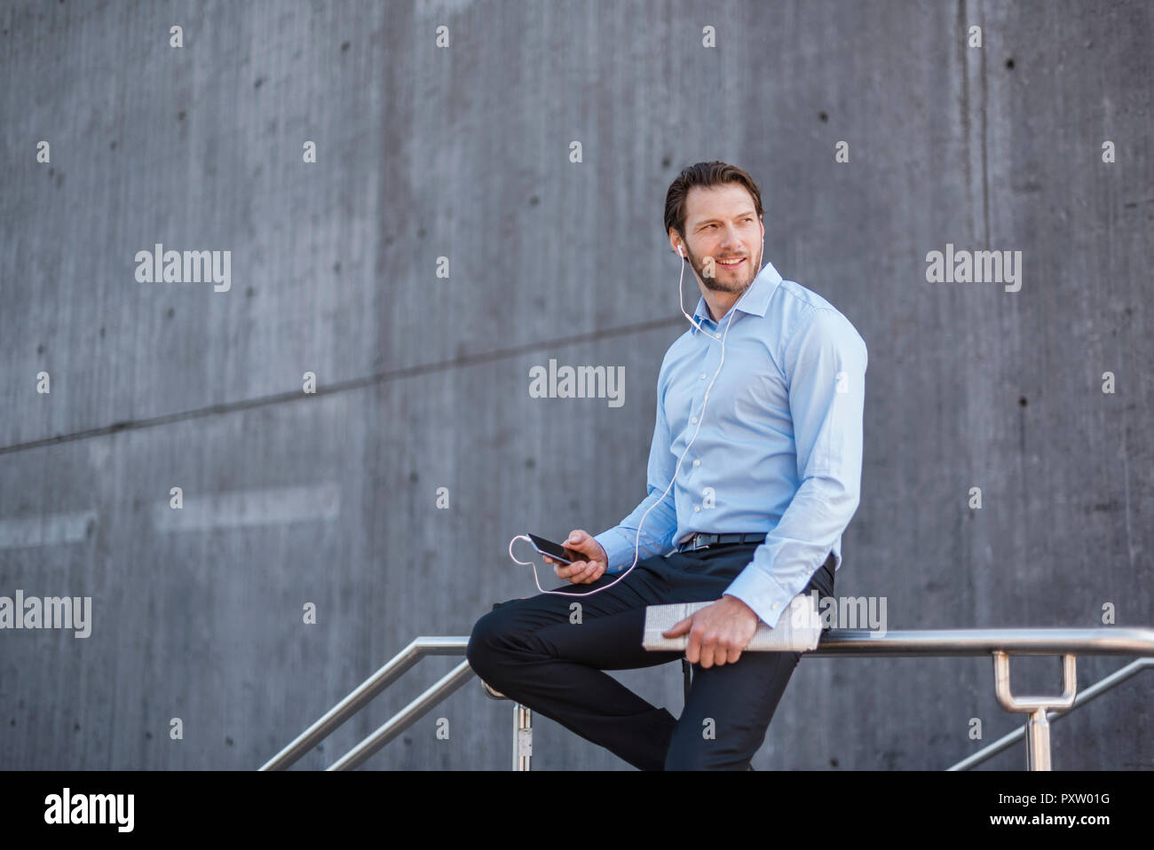 Geschäftsmann mit Smartphone und Ohrhörer sitzen auf einem Geländer Stockfoto