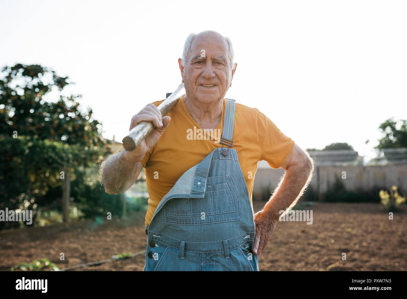 Älterer Mann in Jeans overall Holding Garten Werkzeug auf der Schulter und an der Kamera Stockfoto