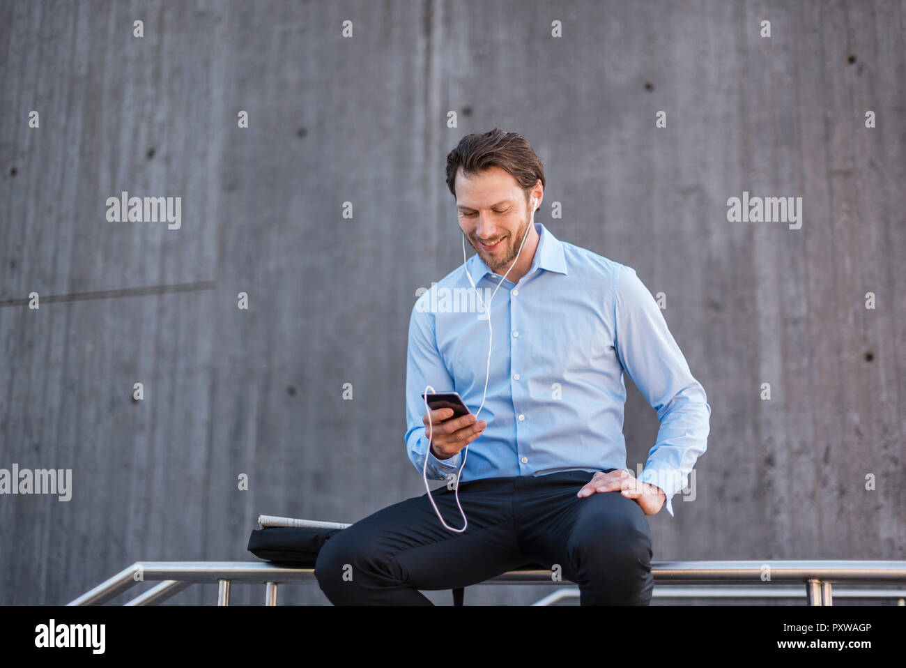 Lächelnd Geschäftsmann mit Ohrhörern sitzen auf einem Geländer am Smartphone suchen Stockfoto