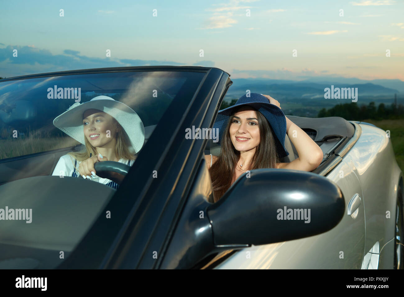 Hübsche Modelle in Dunkelblau und Weiß Rand breite Hüte sitzen auf Vordersitzen von Silber Cabrio. Adorable Mädchen fahren Auto, weg schauen und lächeln. Konzept von luxuriösen Automobil und Reise. Stockfoto