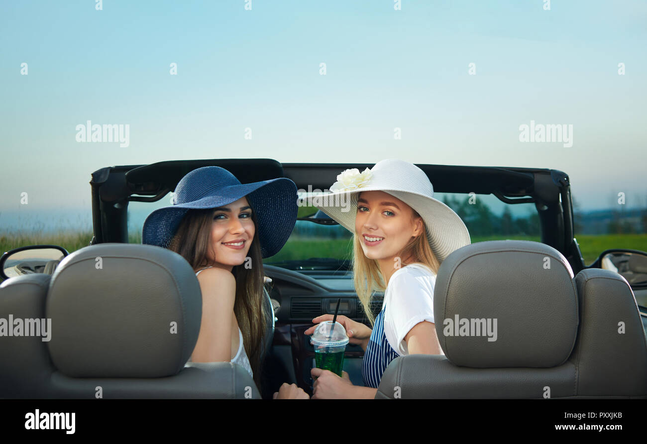 Ansicht von der Rückseite der zwei junge Frauen sitzen auf Vordersitzen von Cabrio. Bezaubernden brünetten und blonden Modelle zurück in die Kamera schauen und lächeln. Mädchen, die in Weiß und Dunkelblau Rand breite Hüte. Stockfoto