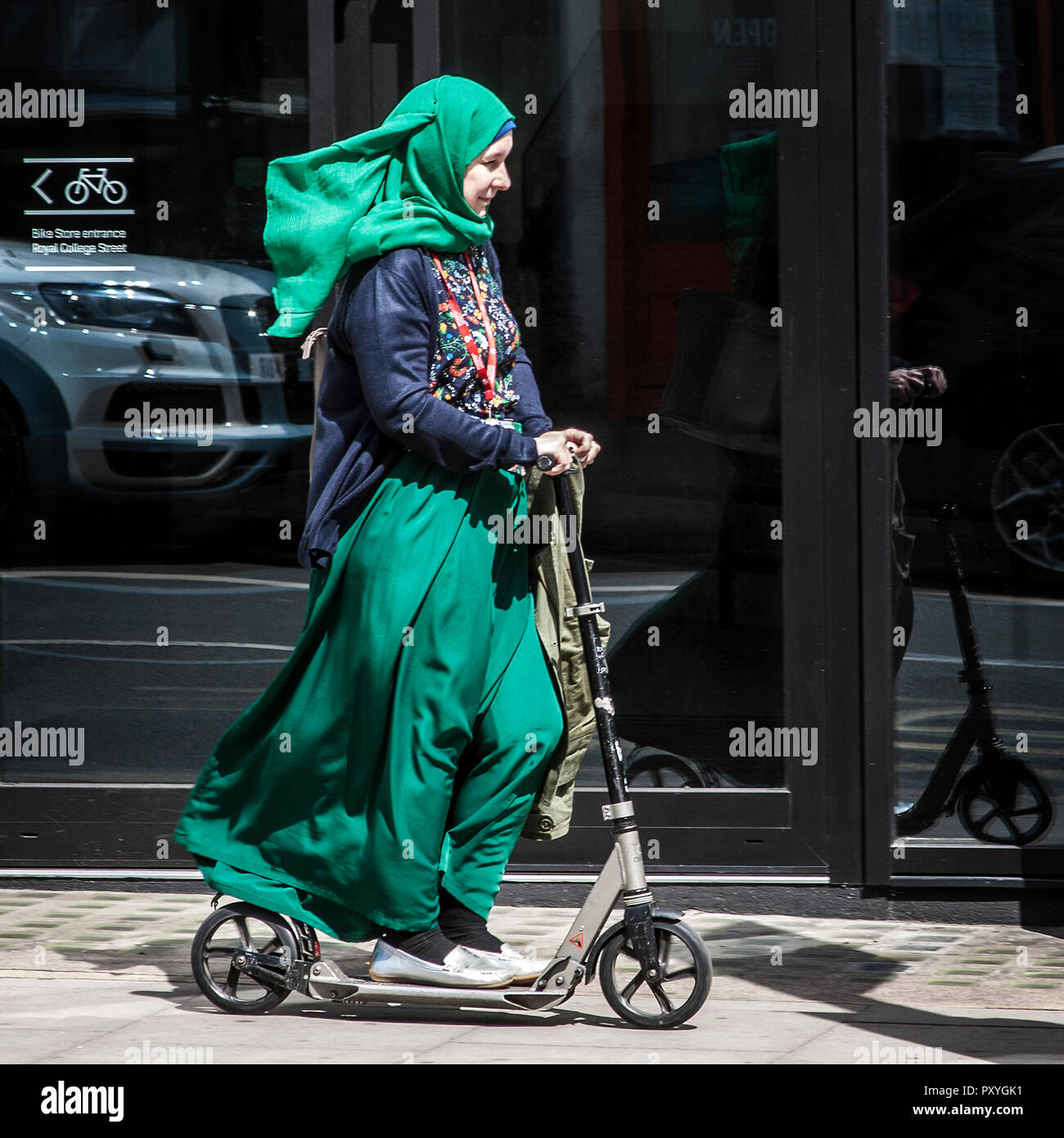 Muslimische Frau im Hidschab-Kick-Roller. Muslimische Frau mit Kopftuch, die auf einem Erwachsenen-Roller auf dem Bürgersteig fährt Stockfoto