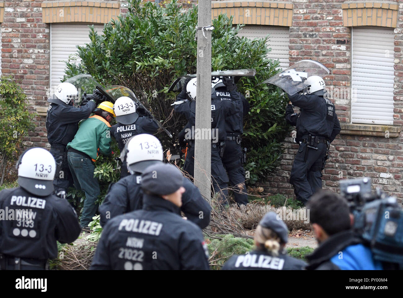 Manheim, Deutschland. 25 Okt, 2018. Polizisten schützen Ein Arbeitnehmer, der aus einem Busch vor einer der besetzten Häusern. Nach der Evakuierung von einem Protest Camp während der Nacht, am Donnerstag morgen den task forces begannen Aktivisten von besetzten Häusern in Kerpen Bezirk zu bringen. Der Protest der Gruppe, die die leerstehenden Gebäude am Rand des Tagebaus seit letzter Woche besetzt hatte, wollte nicht, dass die Häuser freiwillig zu verlassen, sagte die Polizei. Das Dorf Manheim, um Wege zu öffnen. Geben Tagebau Credit: Henning Kaiser/dpa/Alamy leben Nachrichten Stockfoto