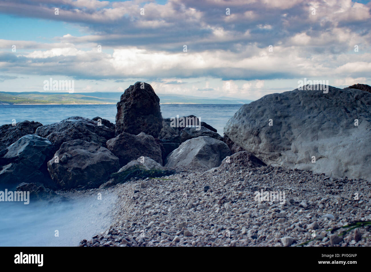 Surf Wellen des Meeres in Kroatien mit Steinen am Strand. Wellen gegen die Felsen und gesprüht. Im Hintergrund sind dunkle Wolken am blauen Himmel. Stockfoto