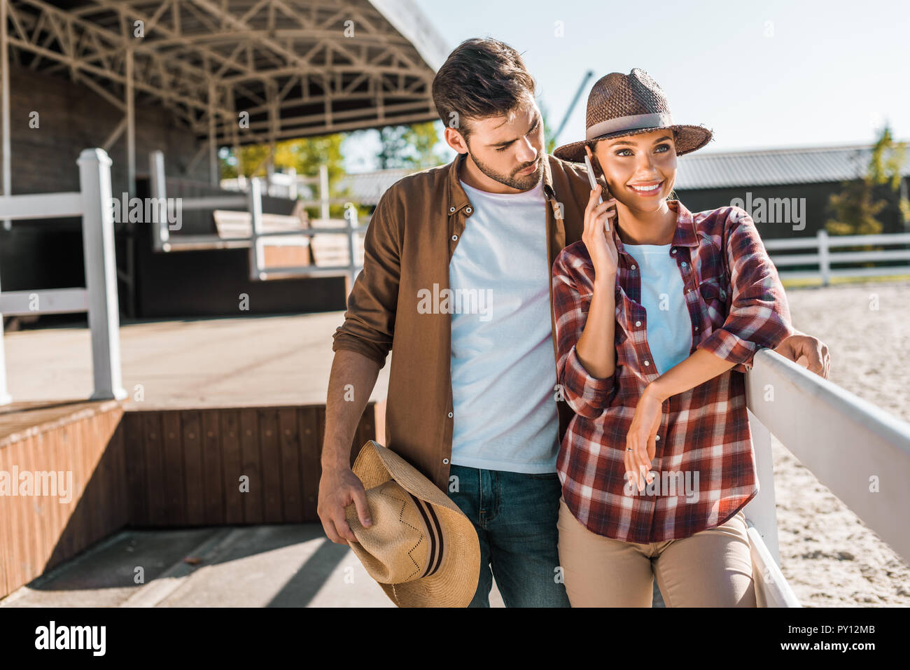 Stilvolle Cowboy und Cowgirl in Freizeitkleidung in der Nähe von Zaun an der Ranch stehen, Frau sprechen von Smartphone Stockfoto