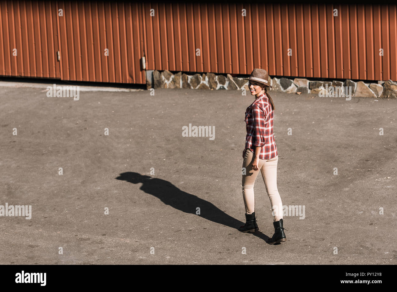 Lächelnd attraktive Cowgirl in kariertem Hemd und Hut zu Fuß in der Nähe der braunen Gebäude an der Ranch Stockfoto