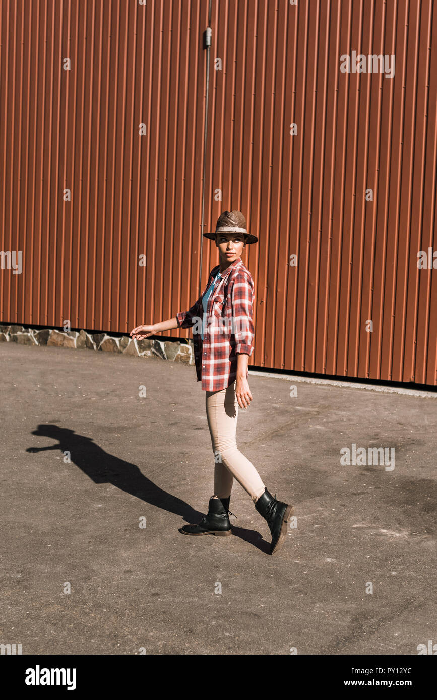 Attraktive cowgirl in kariertem Hemd und Hut zu Fuß in der Nähe der braunen Gebäude an der Ranch Stockfoto