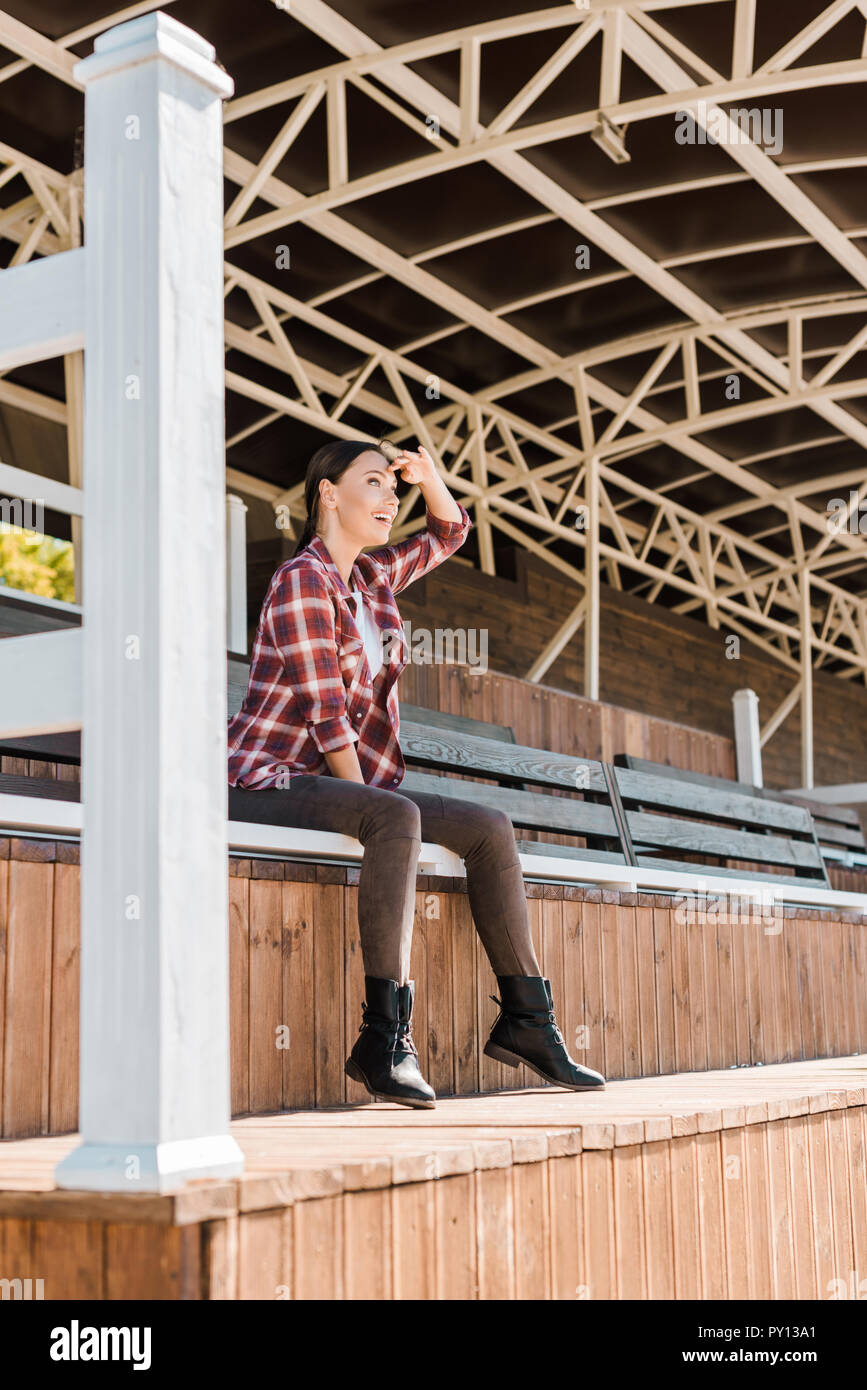 Attraktive cowgirl in kariertem Hemd sitzt auf der Bank an der Ranch Stadion und suchen nach etwas Stockfoto