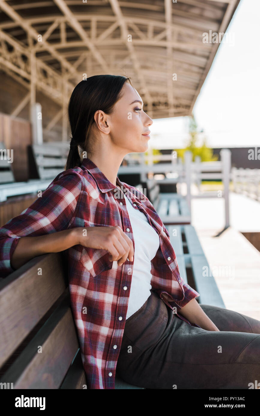 Seitenansicht des schönen Cowgirl in kariertem Hemd sitzt auf der Bank an der Ranch Stadion und Wegsehen Stockfoto