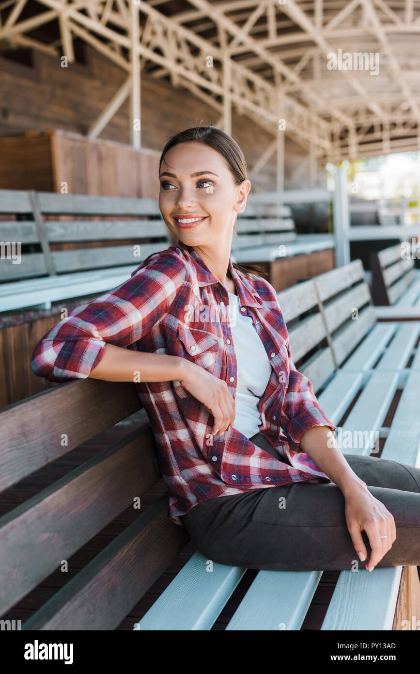 Lächelnd attraktive Frau in kariertem Hemd sitzt auf der Bank an der Ranch Stadion und Wegsehen Stockfoto