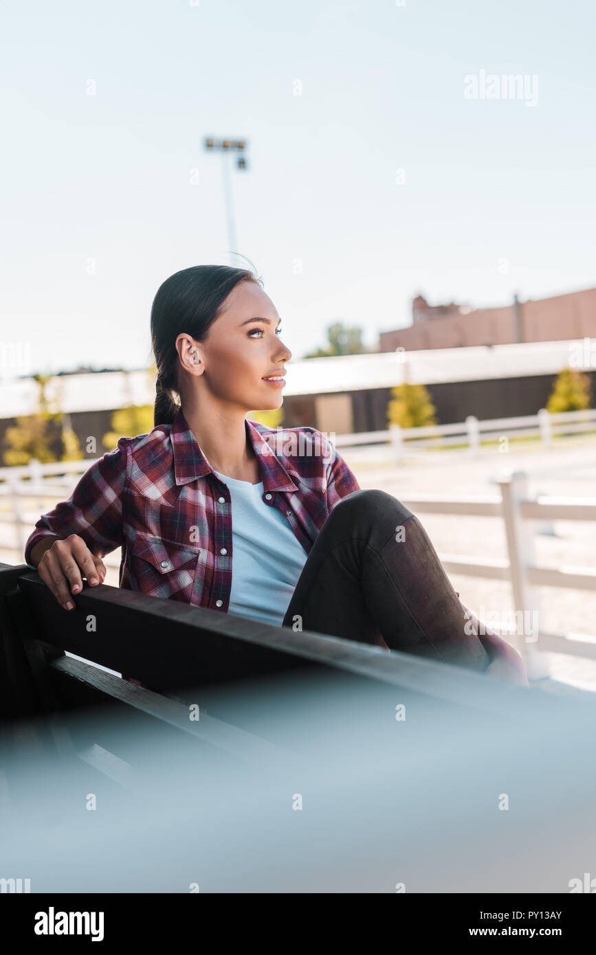 Seitenansicht des attraktiven Cowgirl in kariertem Hemd sitzt auf der Bank an der Ranch Stadion und Wegsehen Stockfoto