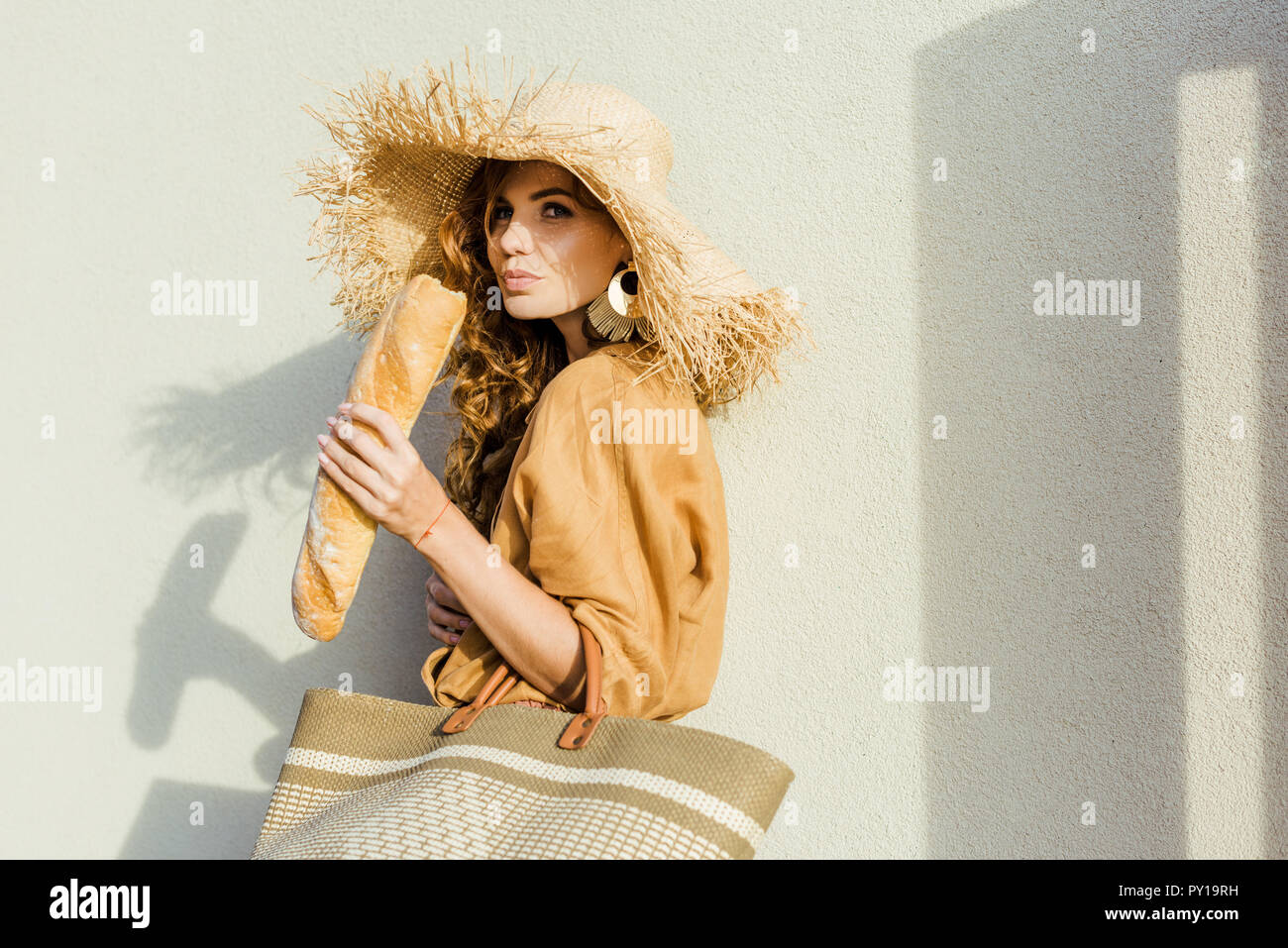 Stilvolle junge Frau mit Strohhut stand vor der weißen Wand und essen Baguette Stockfoto