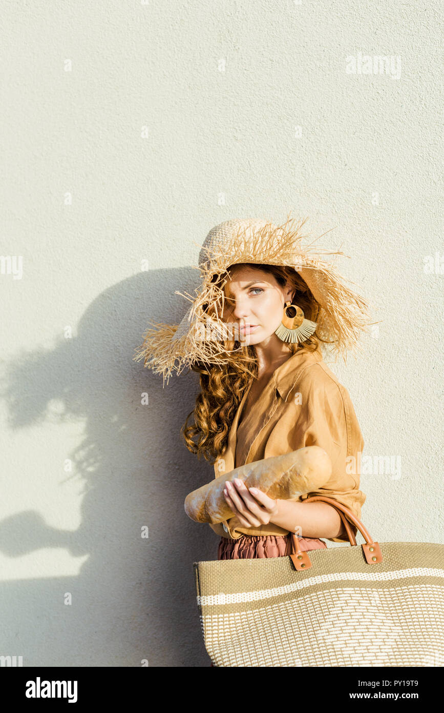 Modische junge Frau im Strohhut mit Baguette stand vor der weißen Wand Stockfoto