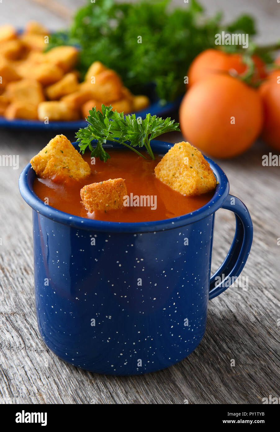 Nahaufnahme von einem blauen Becher mit frischen, hausgemachten Tomatensuppe gefüllt. Tomaten, Petersilie und Croutons unscharf im Hintergrund. Stockfoto