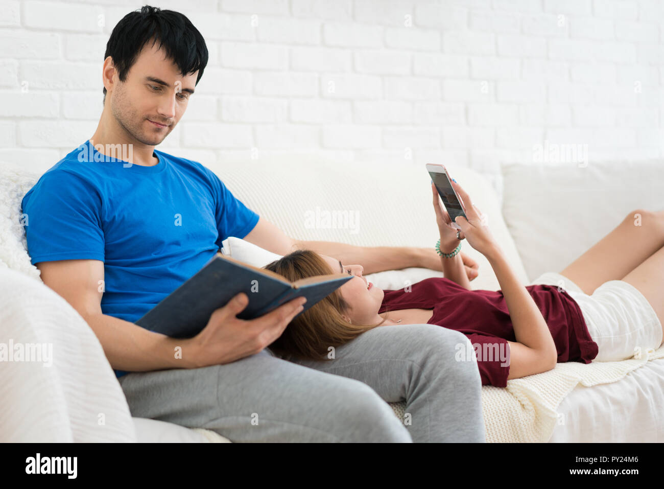 Junges Paar auf dem Sofa entspannen in Ihrem Wohnzimmer zu Hause. Mit Smartphone und ein Buch lesen. Stockfoto