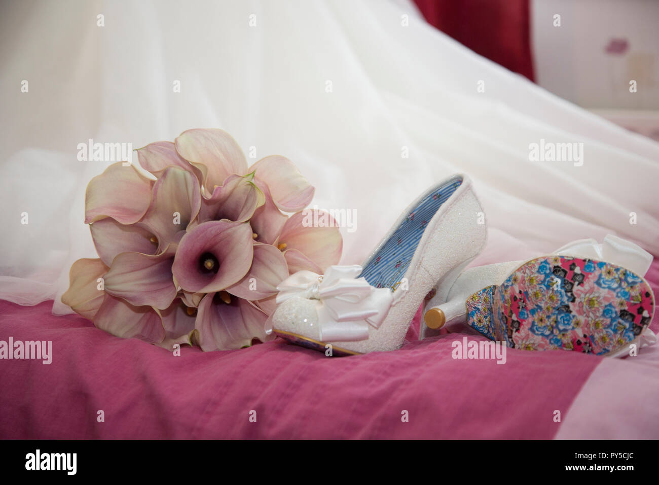 Bräute Schuhe auf dem Bett neben ihr Bouquet von rosa Calla Lilien sitzt am Ende ihrer Hochzeit Kleid Zug wartet an Ihrem Hochzeitstag getragen zu werden. Stockfoto