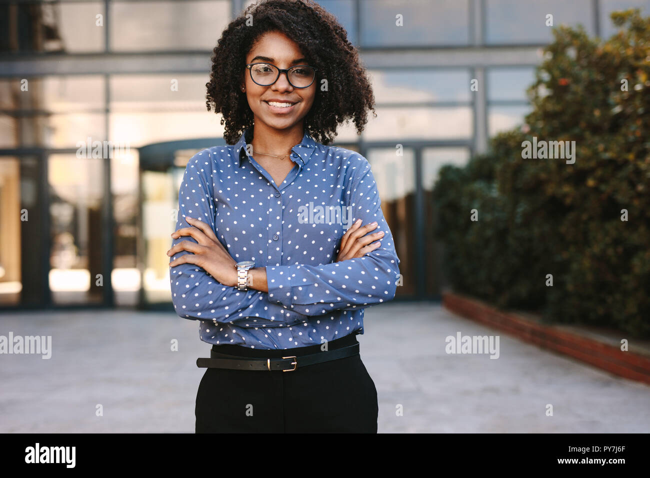 Porträt einer positiven jungen afrikanerin, die draußen mit überkreuzten Armen steht. Geschäftsleute stehen selbstbewusst mit Bürogebäuden in BA Stockfoto