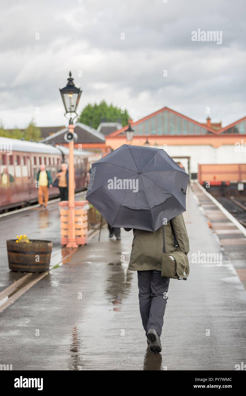 Rückansicht einer trainspotter-Frau in Anorak, die den Regenschirm hält, der im Regen am Bahnsteig des Vintage-Bahnhofs in Großbritannien von der Kamera wegläuft. Stockfoto