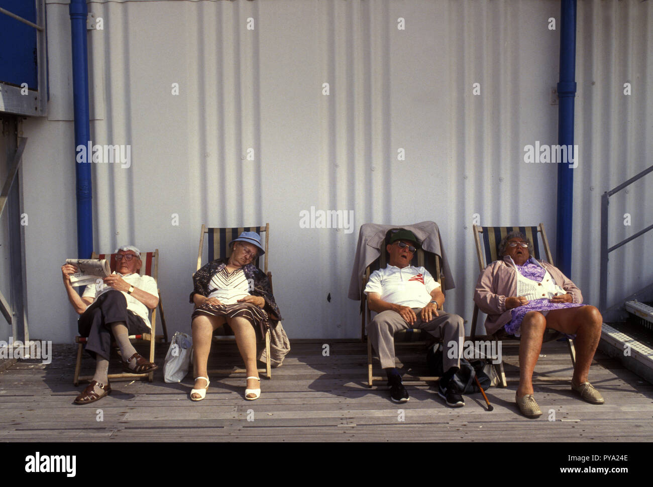 Vier alte Leute sitzen in Liegestühlen, Great Yarmouth. Drei schlafen mit Beinen akimbo, ein Mann sitzt mit gekreuzten Beinen und liest eine Zeitung Stockfoto