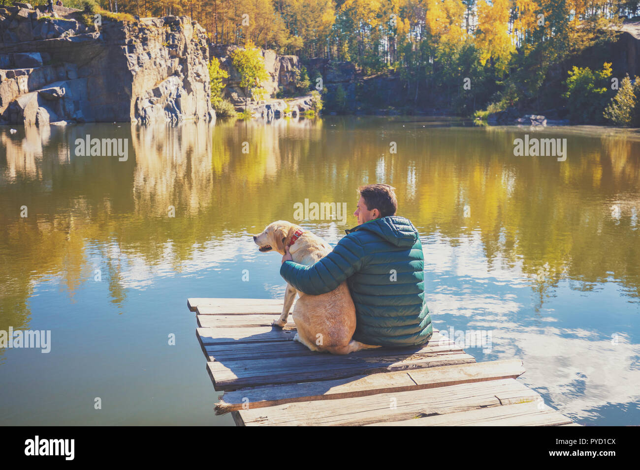 Ein Mann mit Labrador Retriever Hund sitzt auf der Holzterrasse auf der schönen Granite Lake Shore und Suchen am Wasser Stockfoto
