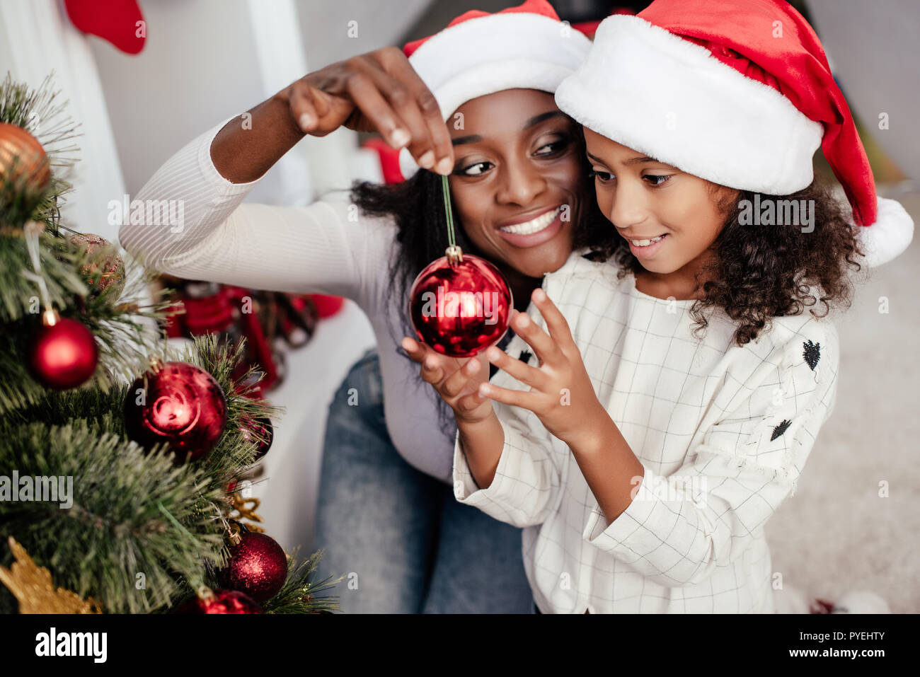 Lächelnd afrikanische amerikanische Frau in Santa Claus hat hilft Tochter Weihnachtsbaum zu Hause zu verzieren Stockfoto