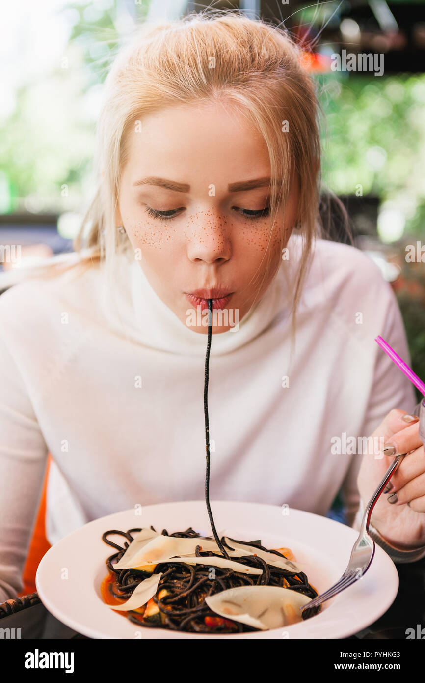 Junge schöne Frau essen schwarze Nudeln mit Meeresfrüchten und Tintenfisch Tinte in das Restaurant im Freien. Lustig und schön. Stockfoto