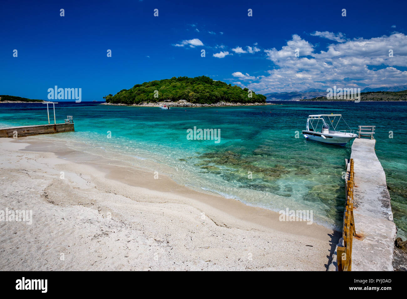 Feder tagsüber schönen Ionischen Meer mit klarem, türkisfarbenem Wasser, Holz- pier und feinen Sand der Küste Blick von Ksamil Strand, Albanien. Tief blauen Himmel mit weißen Wolken. Stockfoto