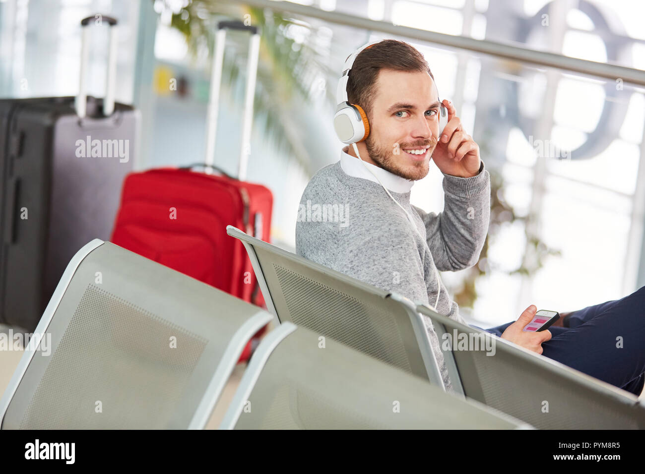 Mann im Flughafen wartet auf seinen Flug und hört Musik mit Kopfhörern und Smartphone Stockfoto