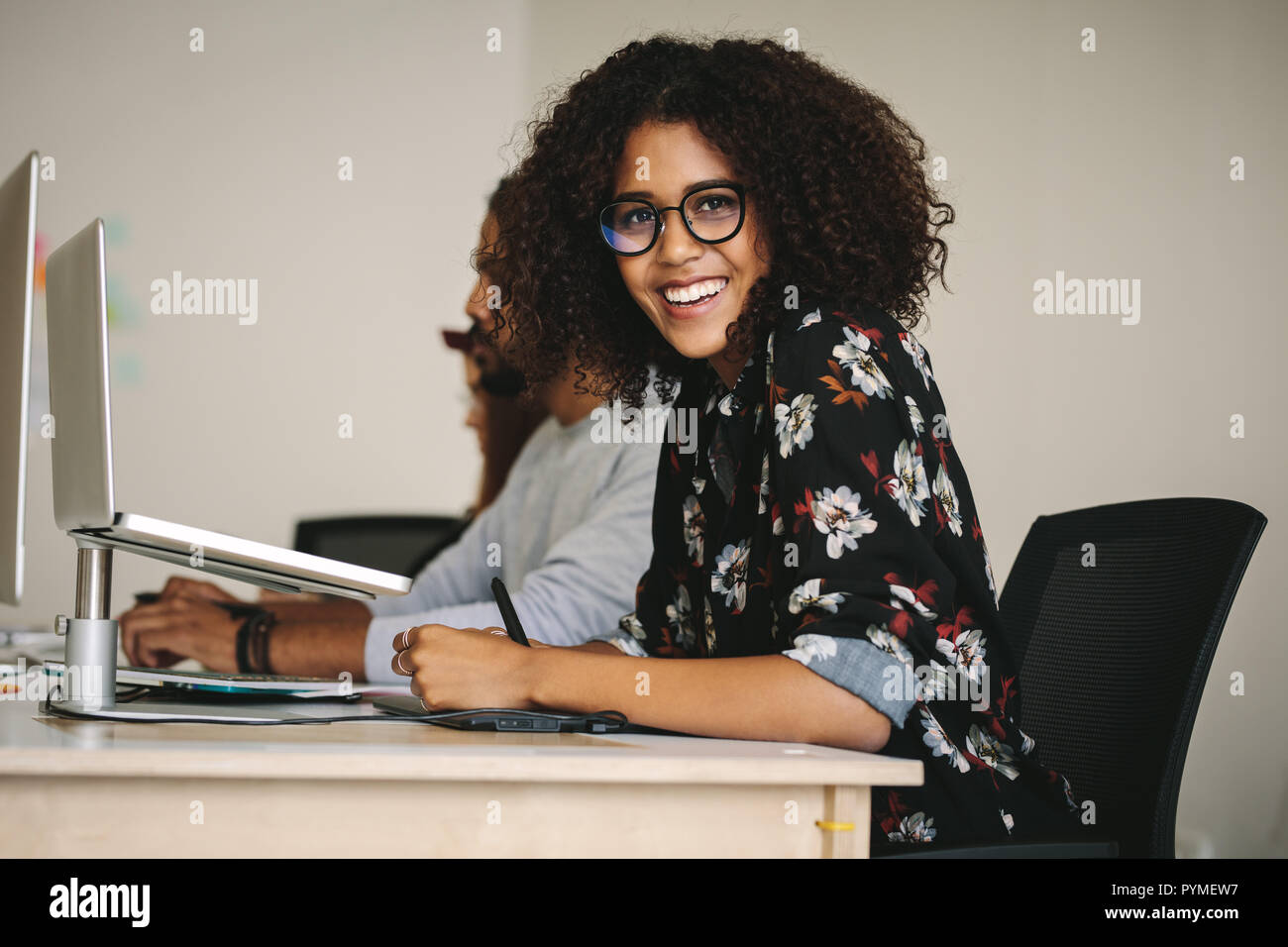 Geschäftsleute, die an ihren Arbeitsplätzen im Büro sitzen. Lächelnde Geschäftsfrau mit Brille, die an ihrem Schreibtisch sitzt und einen Laptop auf dem Tisch hat. Stockfoto