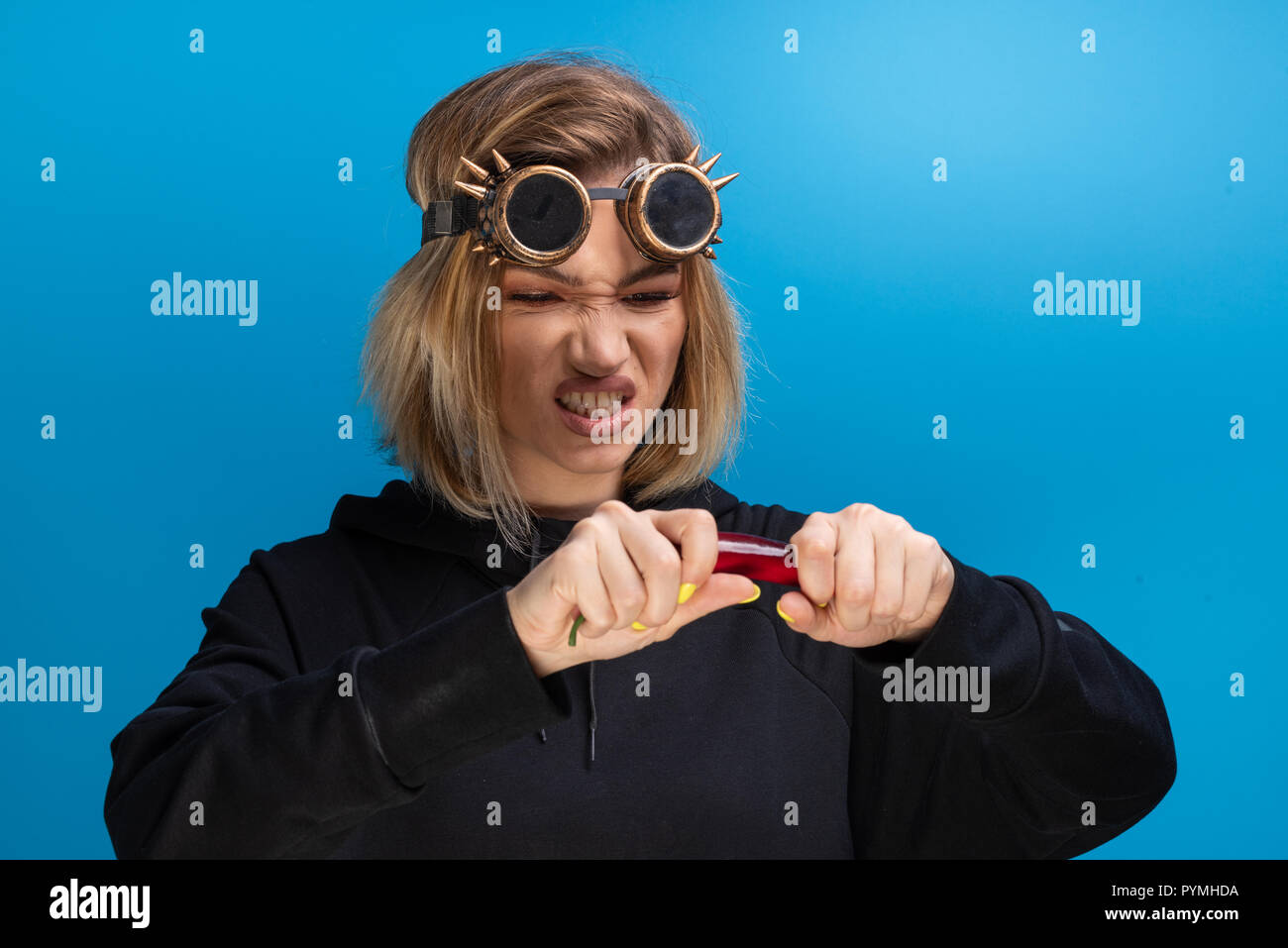Mädchen mit Steam Punk Brille reißen eine rote Chili, während sie wütend. Portraitfotos im Studio vor blauem Hintergrund Stockfoto