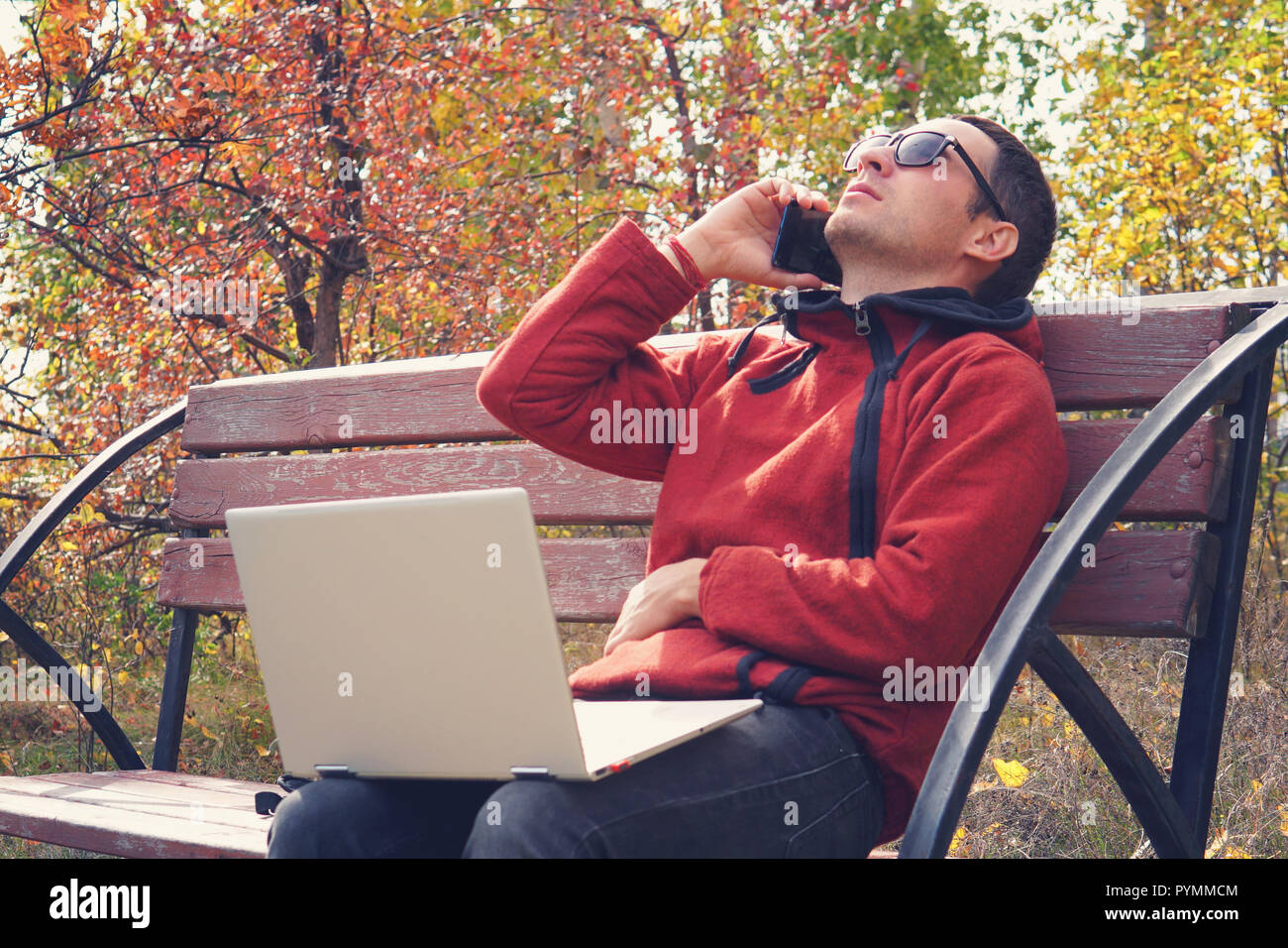 Beschäftigte Person arbeitet online außerhalb der im Urlaub. junger Mann Arbeiten am Laptop Computer in legere Kleidung. Auf dem Smartphone sprechen, sitzt im Park. s Stockfoto