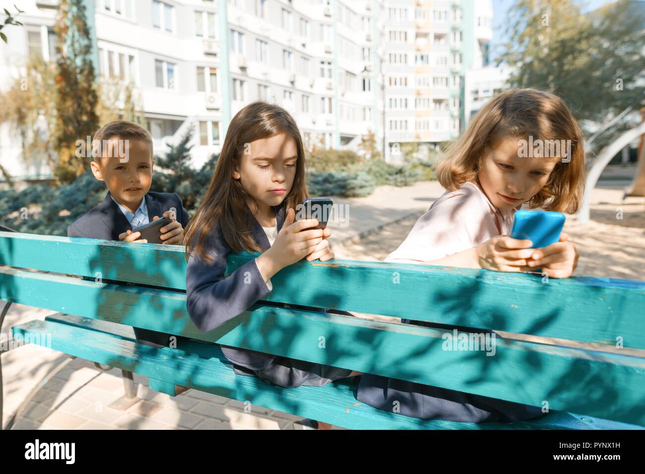 Gruppe von Kindern 7, 8 Jahre alt mit Handys, begeistert, in Smartphones, Schüler mit Rucksäcken, im Hintergrund. Edu Stockfoto