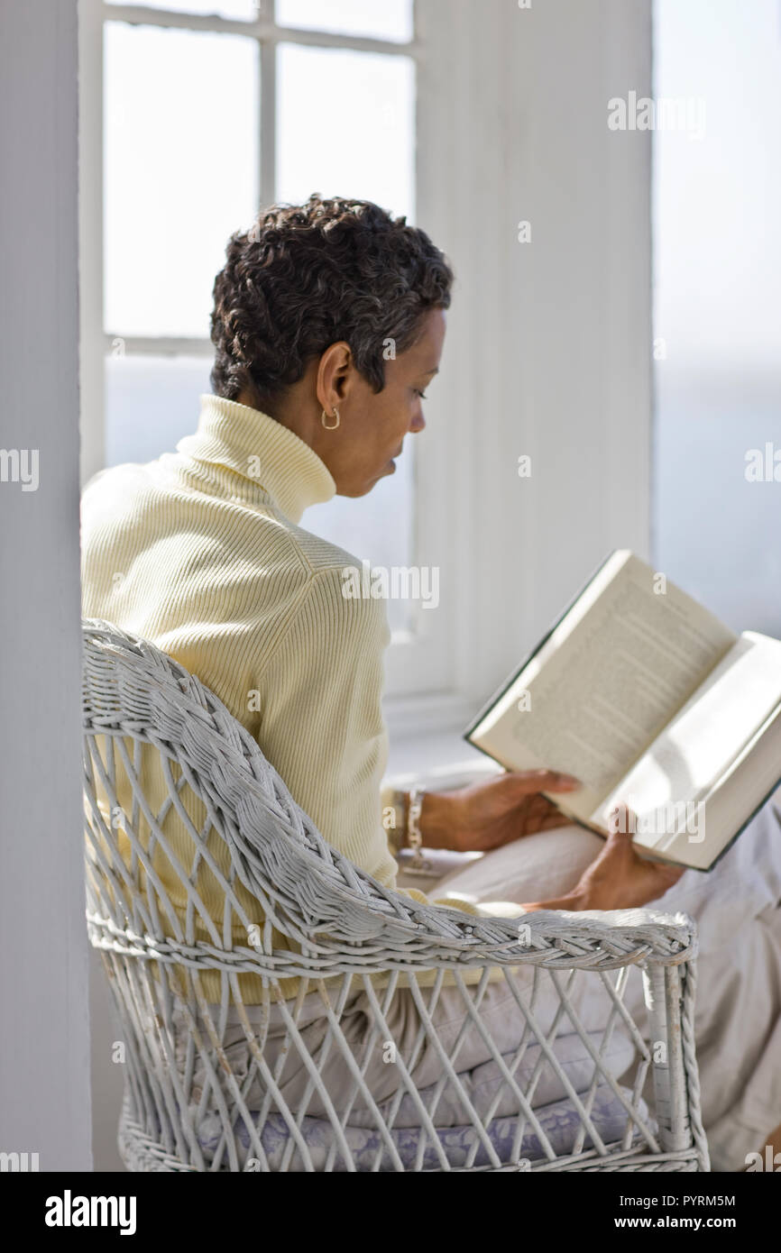 Mitte der erwachsenen Frau in einem korbsessel sitzen, ein Buch zu lesen in einem sonnigen Raum. Stockfoto