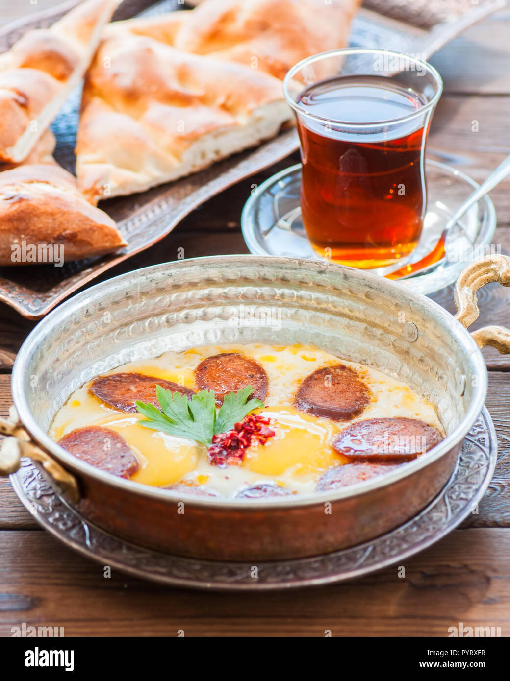 Türkisches Frühstück - Spiegeleier mit Würstchen (sucuk) und Gewürzen in einer Pfanne, Glas Tee auf einem hölzernen Hintergrund. Stockfoto