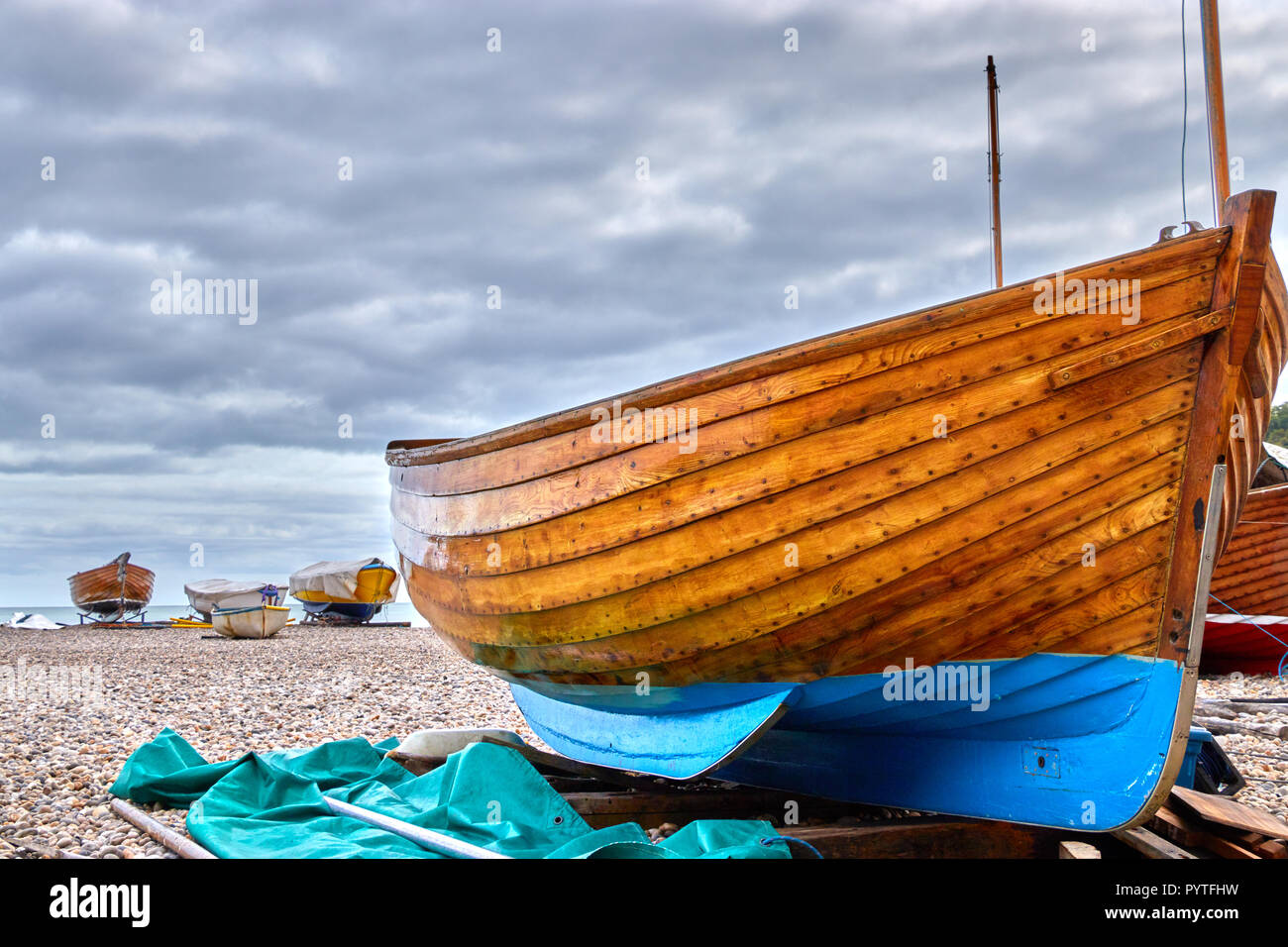 Strände Holzboot auf Bier Strand in Devon, England mit Strände Boote im Hintergrund und Moody bewölkter Himmel Stockfoto