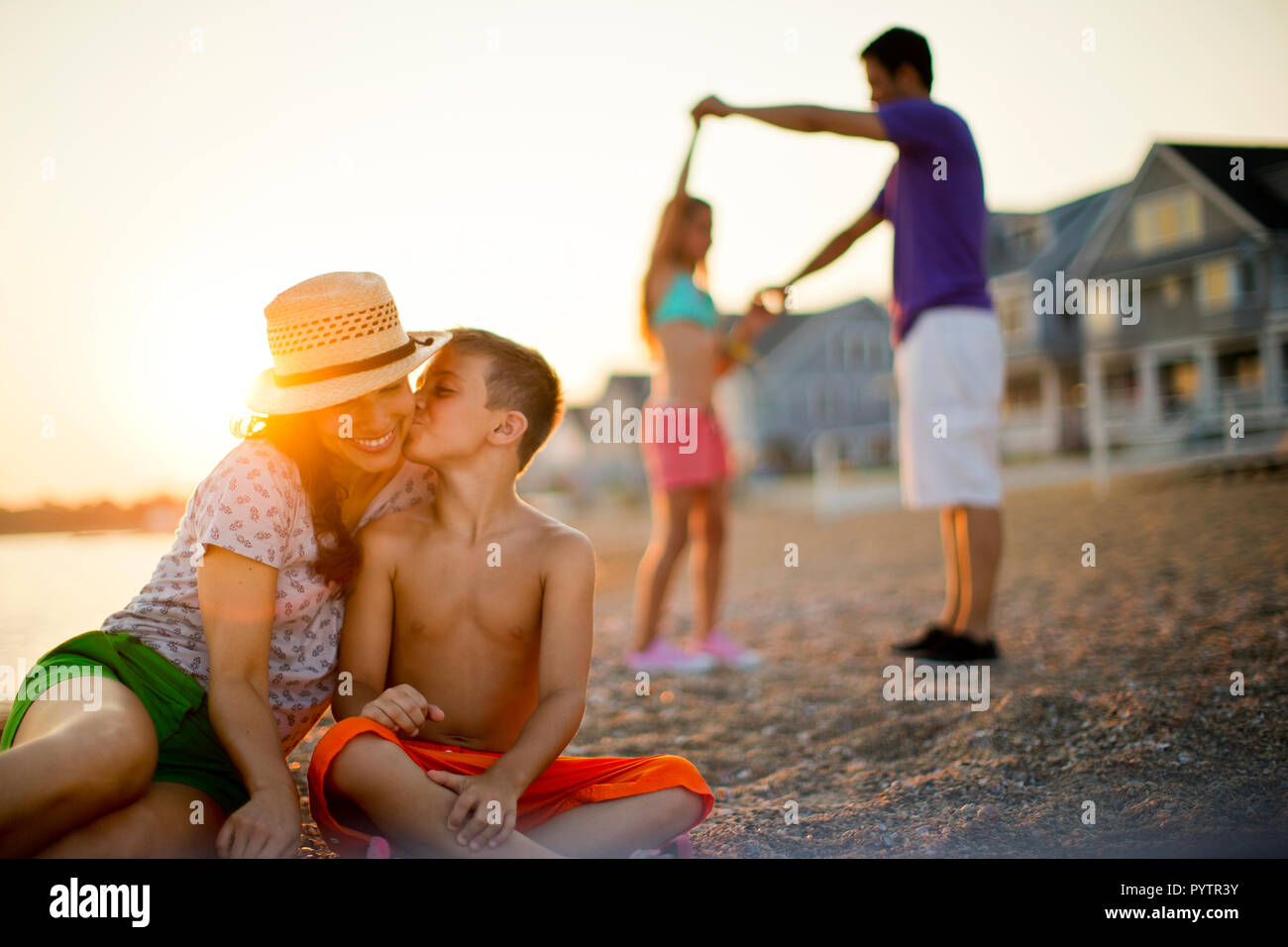 Junge Küsse auf die Wange, während seine Mutter die Familie am Strand bei Sonnenuntergang entspannt. Stockfoto