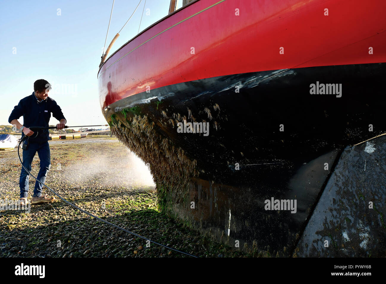 Mann Druck abspritzen übermäßige Algen Unkraut aus den Rumpf seines X Boot, Madame x) wie Es ruht gegen Itchenor's schrubben Delphine Stockfoto