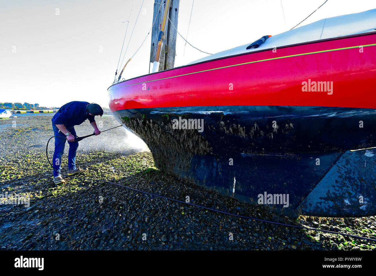 Mann Druck abspritzen übermäßige Algen Unkraut aus den Rumpf seines X Boot, Madame x) wie Es ruht gegen Itchenor's schrubben Delphine Stockfoto