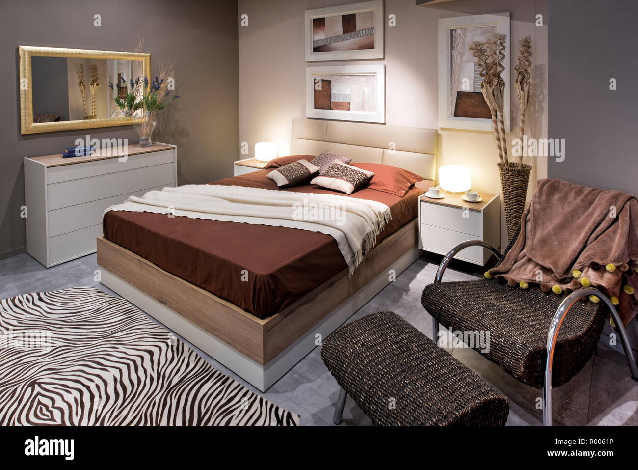 Schlafzimmer mit Doppelbett Gestaltung mit hohen braunen Bett und komfortable Sessel mit Ottomane, zebra Teppich auf dem Boden und Gemälden an den Wänden Stockfoto