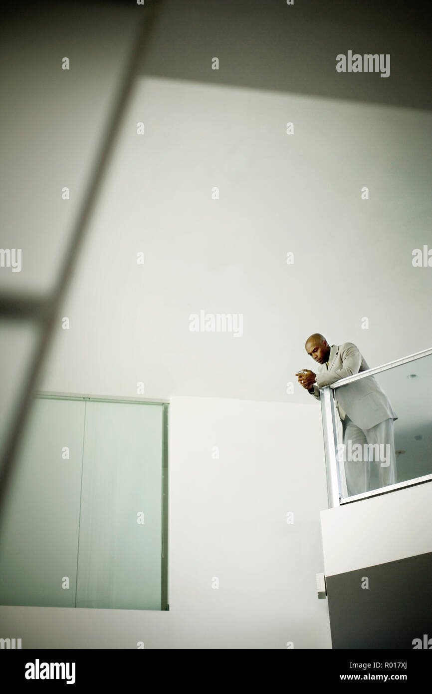Mitte - Erwachsene, Geschäftsmann, lehnend auf einem Geländer, während Sms auf seinem Handy in einem Bürogebäude. Stockfoto