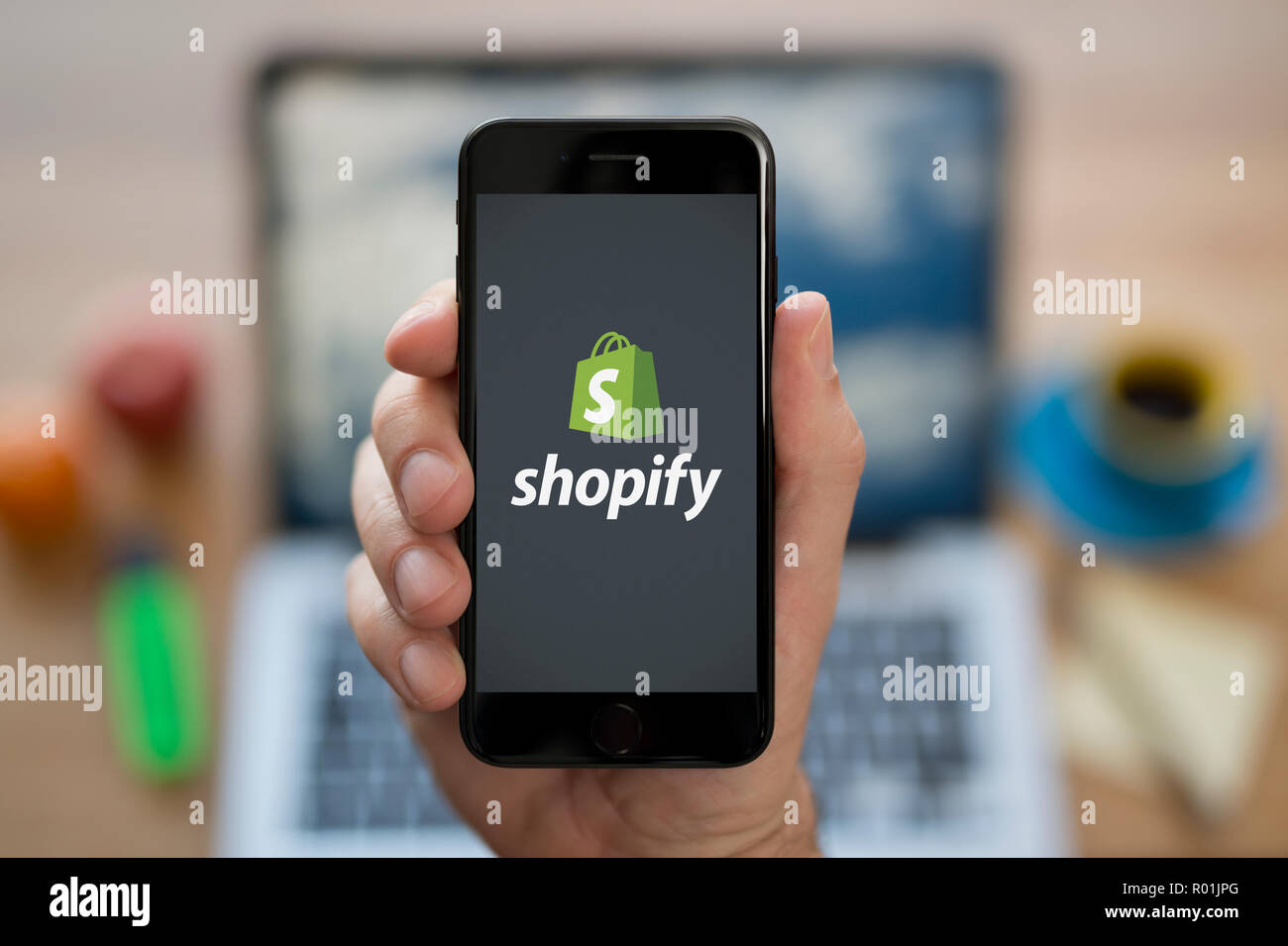 Ein Mann schaut auf seinem iPhone die zeigt die Shopify logo, während saß an seinem Computer Schreibtisch (nur redaktionelle Nutzung). Stockfoto