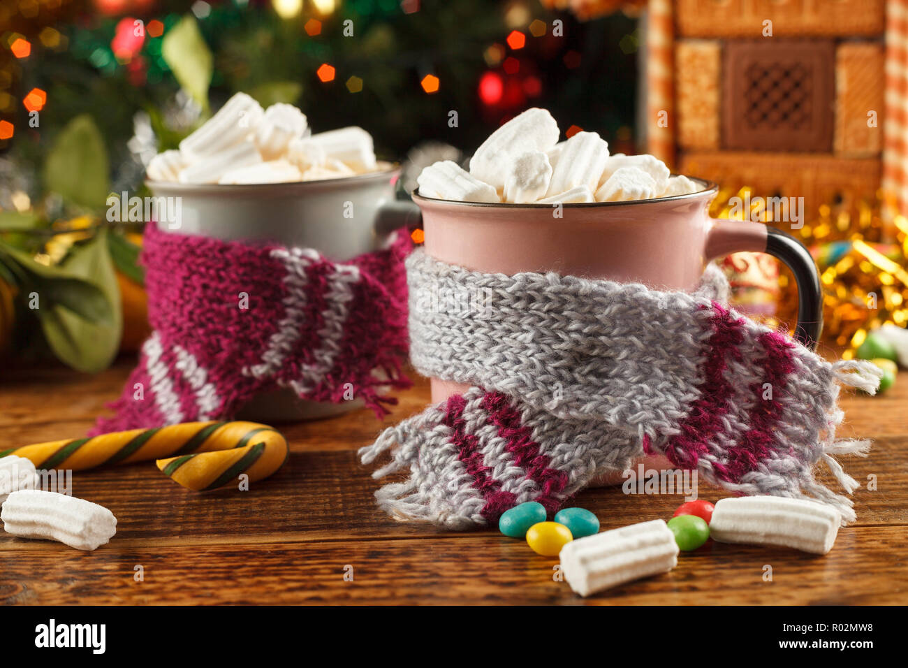 Zwei Tassen in Schals mit einem heißen Getränk und Marshmallows auf dem Hintergrund einer Lebkuchenhaus und einen Weihnachtsbaum. Close-up. Stockfoto
