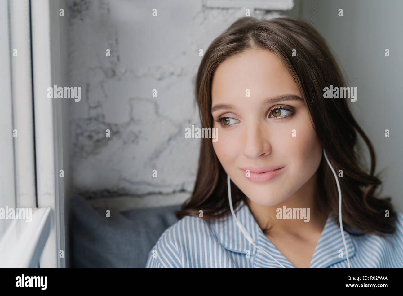 Headshot von attraktiven weiblichen meloman genießt Musik im Kopfhörer mit hoher Lautstärke, friedliche Atmosphäre mag, schaut nachdenklich am Fenster, hört fav Stockfoto