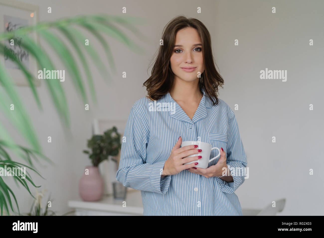 Morgen Zeit Konzept. Junge hübsche europäische Frau in nightclothes trägt weiße Tasse Kaffee oder Cappuccino, steht drinnen im Wohnzimmer, hat Wochenende Stockfoto