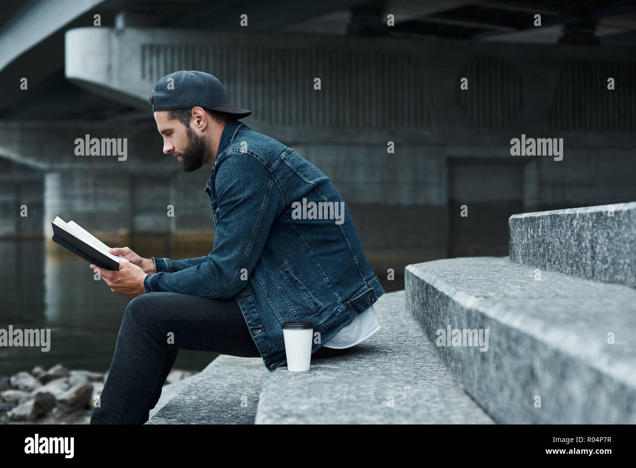 Freizeitaktivitäten im Freien. Junge stilvollen Mann sitzt auf der Treppe auf die Straße der Stadt bei einer Tasse heißen Kaffee beim Lesen Buch fröhliche Seite anzeigen Stockfoto