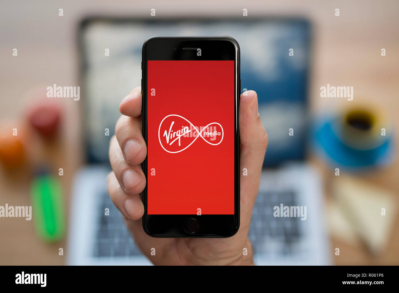 Ein Mann schaut auf seinem iPhone die zeigt die Virgin Media Logo, während saß an seinem Computer Schreibtisch (nur redaktionelle Nutzung). Stockfoto