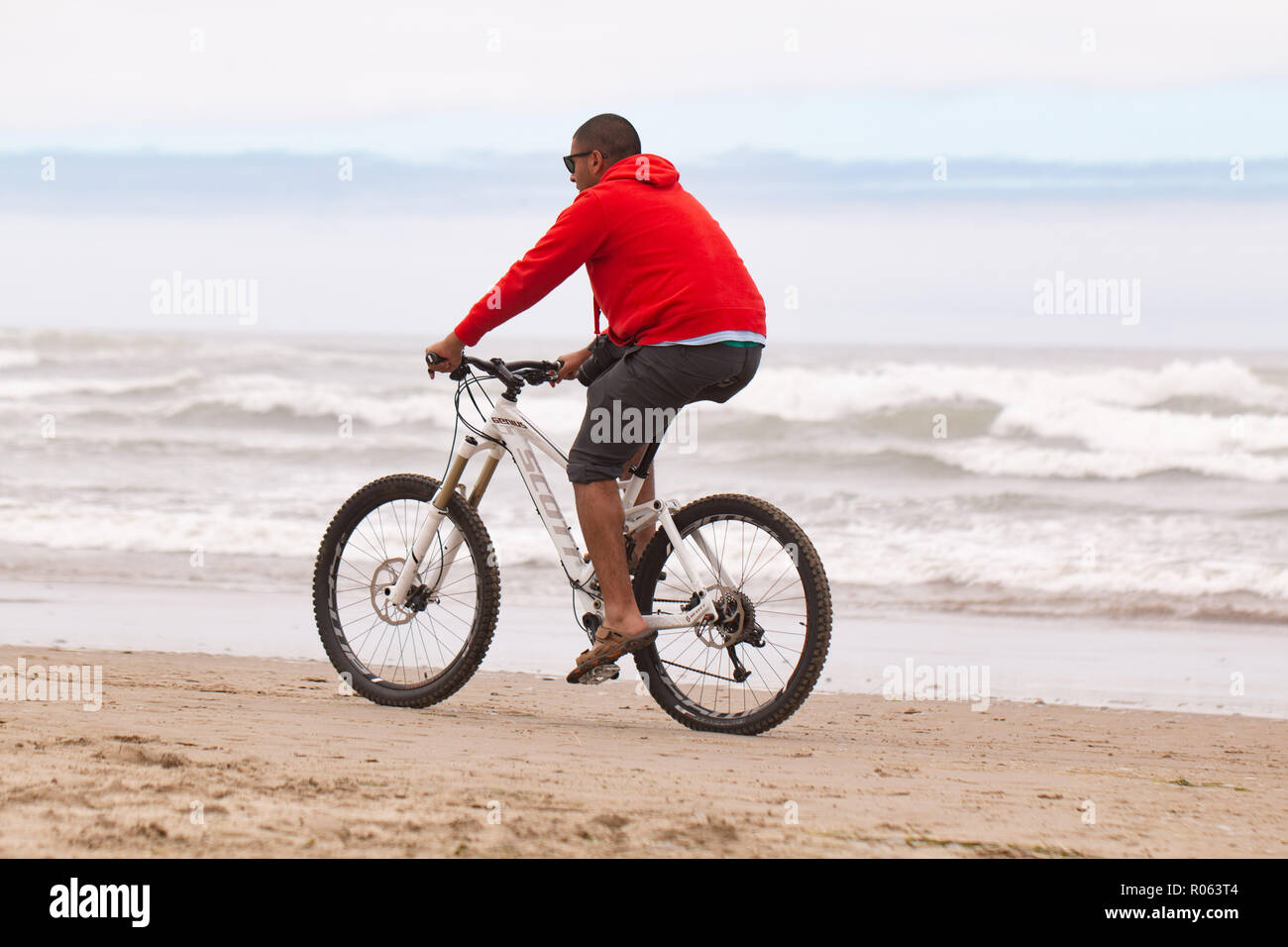 Cannon Beach oder/USA, 2. Juli 2016: der Mann, der in den roten Pullover mit dem Fahrrad entlang des Pazifischen Ozeans Oregon beach Küste auf Sand. Wellen planschen in backgrou Stockfoto