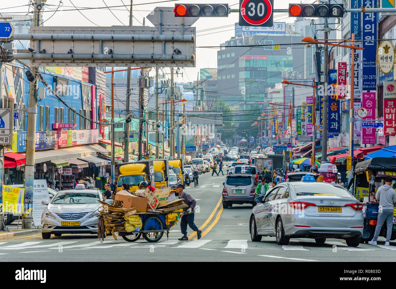 Ein Mann drängt eine Hand Warenkorb geladen mit recyceltem Karton über einen Fußgängerüberweg über eine befahrene Straße in Busan, Südkorea. Stockfoto